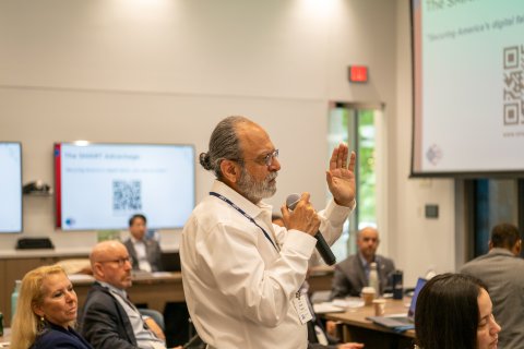 Haresh Malkani of CESMII speaks into a microphone in a large meeting room full of people