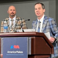 Photo of two men wearing plaid suits standing at a podium with a sign with America Makes' logo on it.