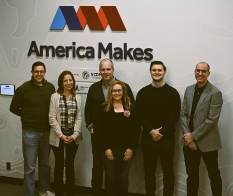 Photo of Brandon Ribic with colleagues in front of the America Makes logo