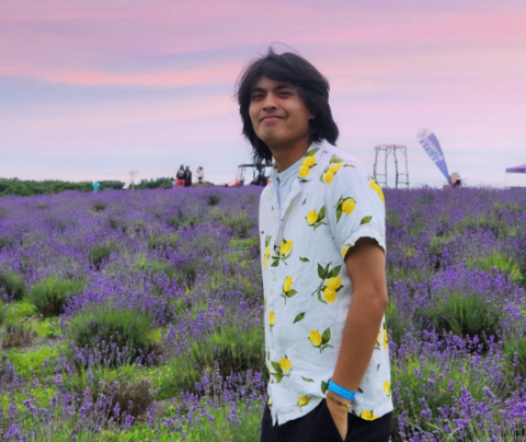 Photo of Alex Nop in a tropical shirt standing in a field of purple flowers with a pink sunset sky behind him.