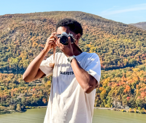 A photo of Keith Joseph taking a picture with his camera. Autumn foliage on a hill behind him.