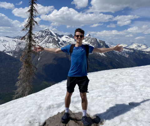 Photo of Ian Goodine on a hike standing on a snow-covered mountain wearing shorts and a tee shirt